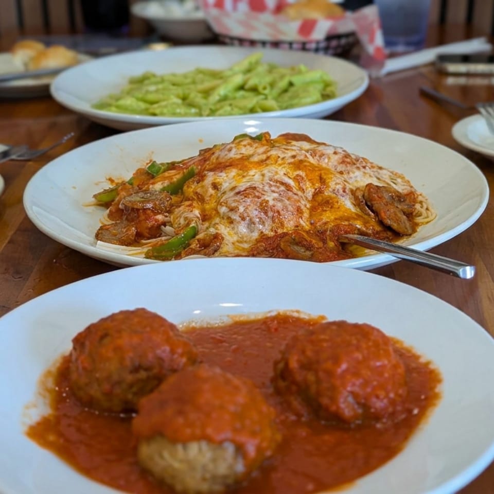 Plates of Italian food, including meatballs in tomato sauce, cheese-covered pasta, and green pasta, arranged on a wooden dining table.