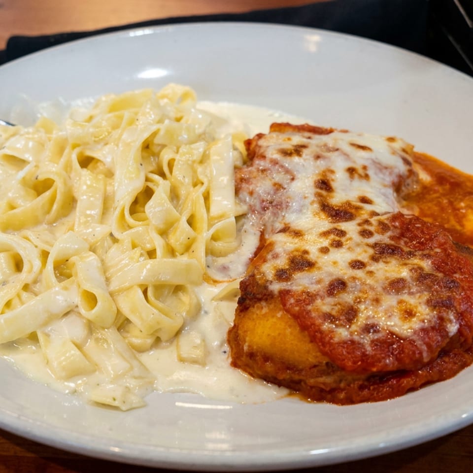 Chicken Parmesan with melted cheese and marinara sauce served alongside creamy fettuccine Alfredo on a white plate.