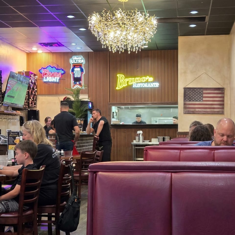 Busy Brumano's Italian restaurant dining room with red booths, chandeliers, diners, televisions, and illuminated beer signs.